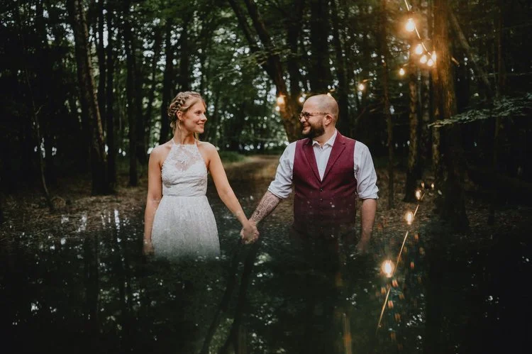 A dreamy and atmospheric woodland wedding portrait captured by Matt Fox Photography, showing a couple standing hand in hand among tall trees and soft glowing string lights. The bride wears a lace halter gown and the groom looks relaxed in a burgundy 