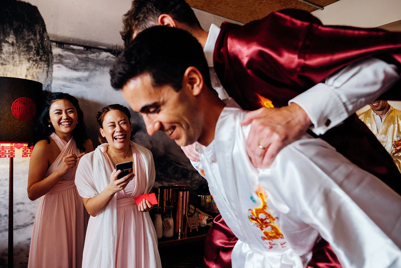 Several people laugh together indoors during a lively moment, with two women in pale pink dresses smiling broadly as they watch two men in embroidered satin robes playfully interacting in the foreground. One man bends forward while another in a red r