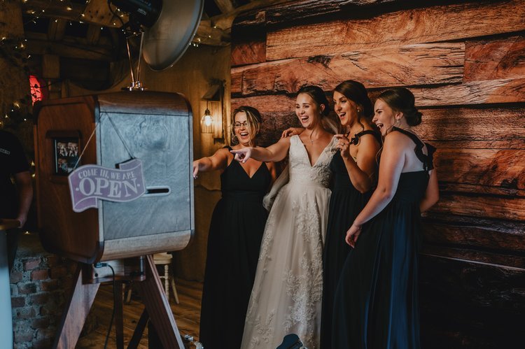 A bride and her bridesmaids share a laugh as they pose in front of the Flash Fox Photo Booth at a rustic barn wedding. The vintage-style booth blends perfectly with the warm wooden setting, capturing the fun and personality of the celebration. Flash 