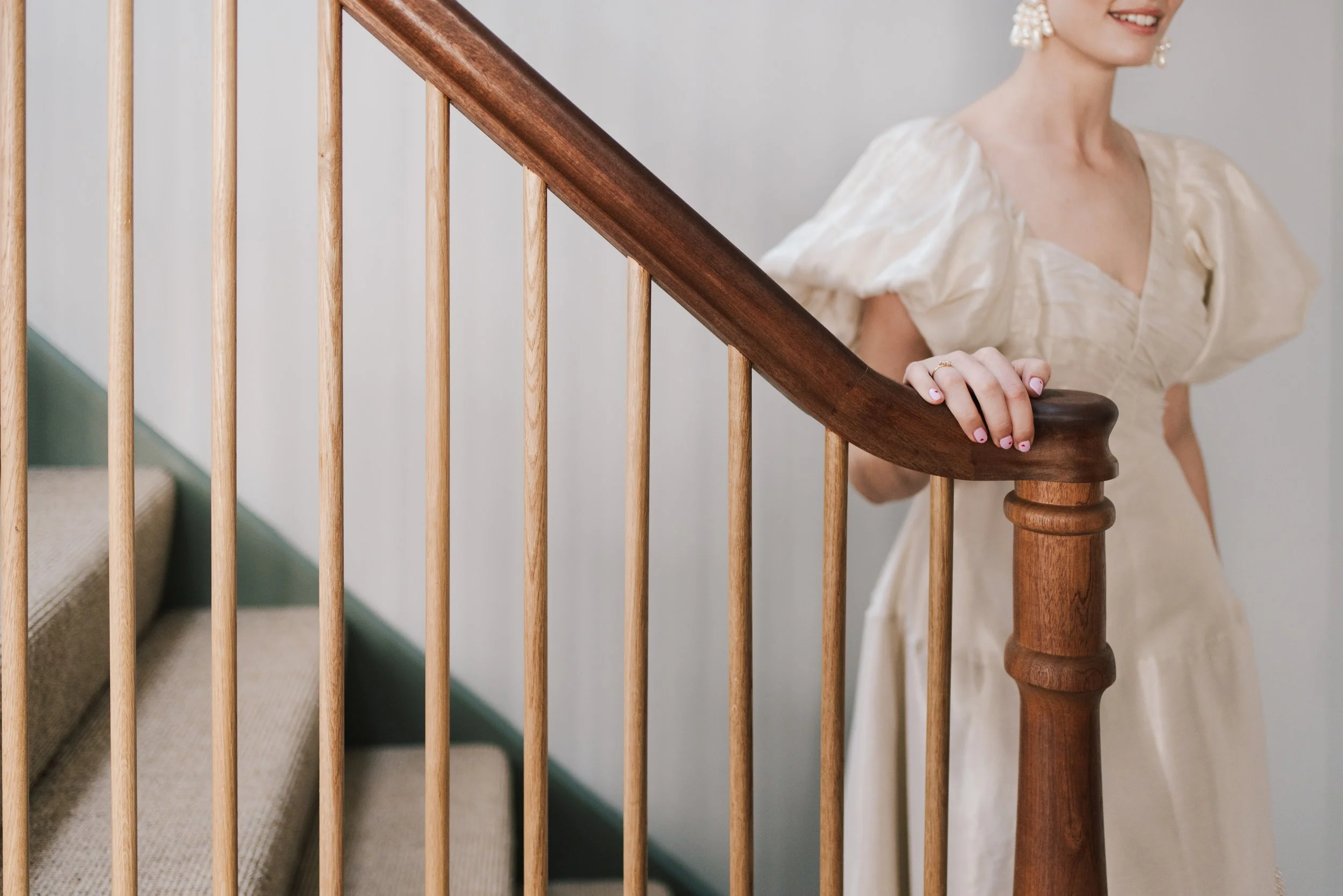 A bride pauses elegantly on a wooden staircase, her hand resting gently on the polished banister to reveal delicate jewellery and beautifully painted nails. The soft puff sleeves and textured fabric of her dress add a romantic, vintage-inspired touch
