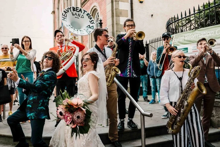 A vibrant and joy filled wedding moment captured by Amy Faith Photography, showing the couple celebrating amongst a lively brass band on the steps of a city street. The bride, in her detailed lace gown and sunglasses, beams with excitement as musicia