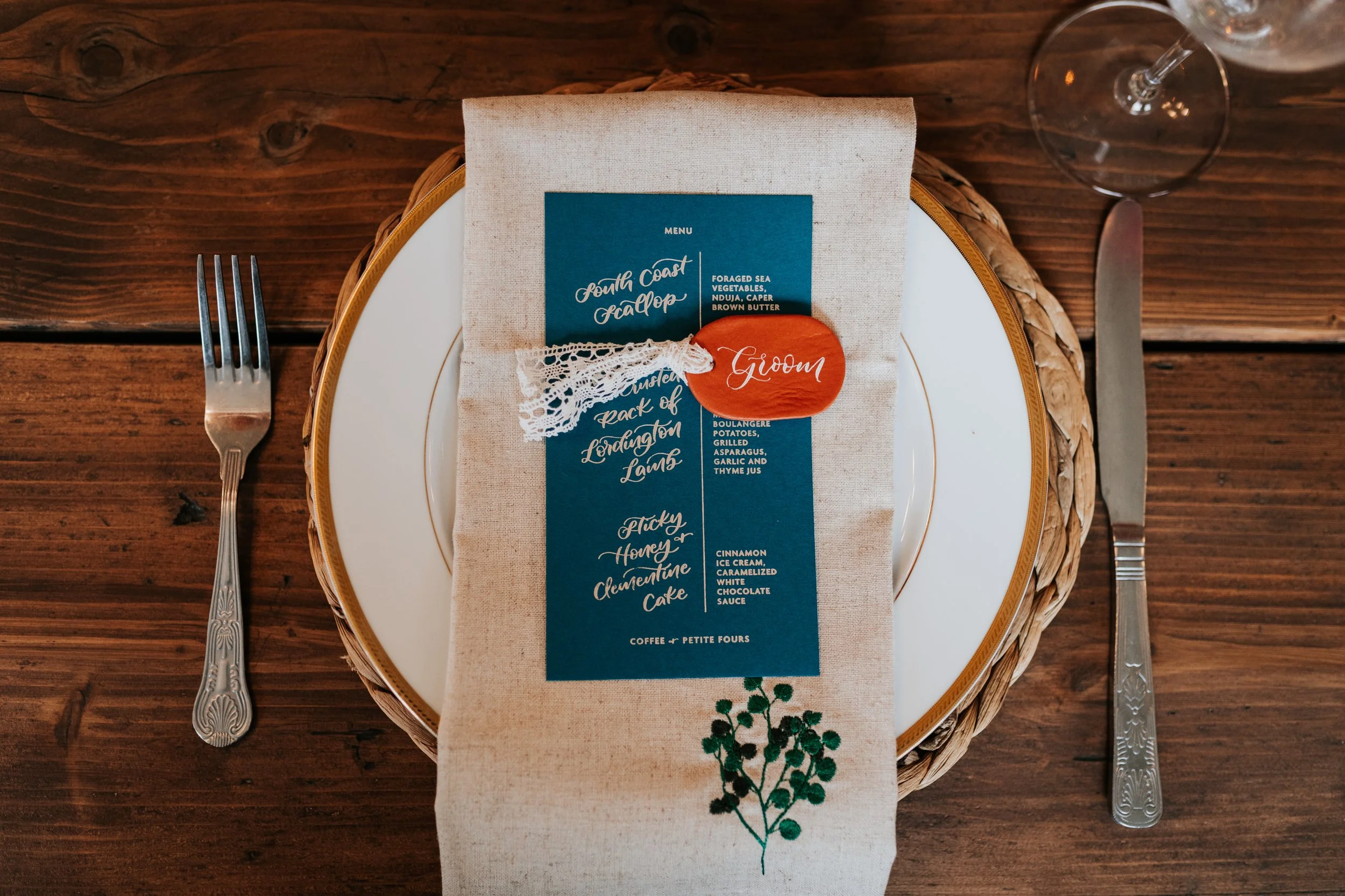 A beautifully styled rustic wedding place setting featuring a woven charger plate, elegant white china, and a natural linen napkin embroidered with greenery. A deep teal wedding menu card sits on top, tied with delicate lace and a hand-painted terrac