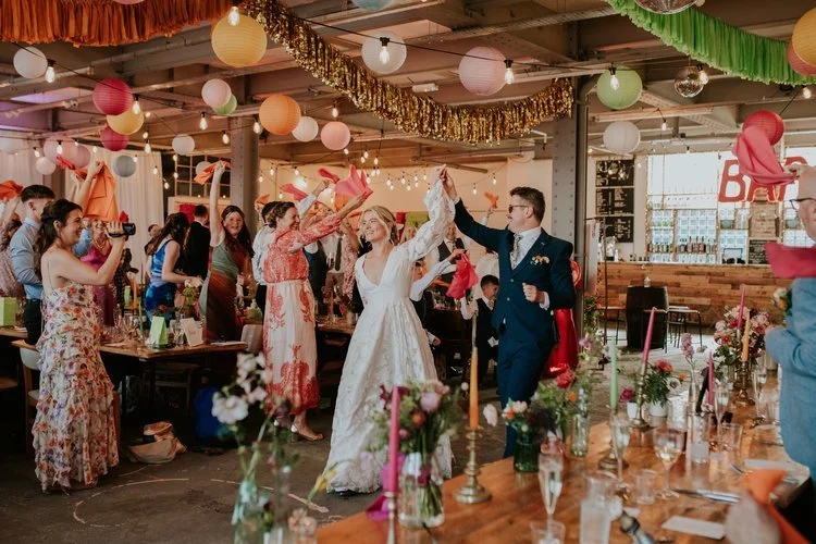 A colourful wedding celebration styled by The Village Green Events, Sheffield. The newlyweds make a joyful entrance surrounded by guests waving napkins under a ceiling of vibrant lanterns, gold tinsel, and string lights. The rustic-industrial venue f
