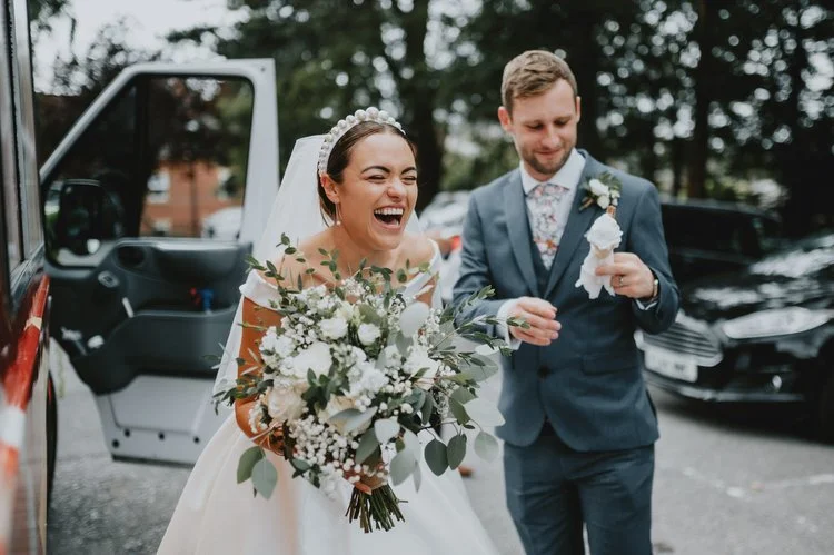A joyful and candid wedding moment captured by Matt Fox Photography, showing the bride laughing wholeheartedly as she holds a lush bouquet of white flowers and greenery. Beside her, the groom smiles while unwrapping a small gift or keepsake, both cle