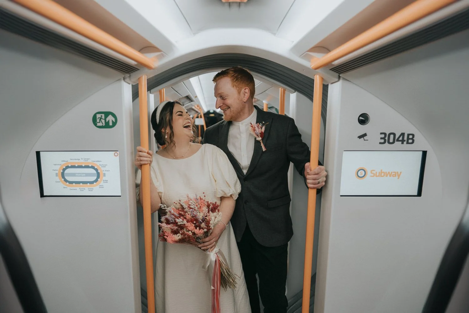 A newlywed couple laugh together inside a modern subway train, the bride holding a dried floral bouquet in a relaxed white wedding dress while the groom wears a dark suit, captured as candid, documentary-style wedding photography by Good Luck Wolf in