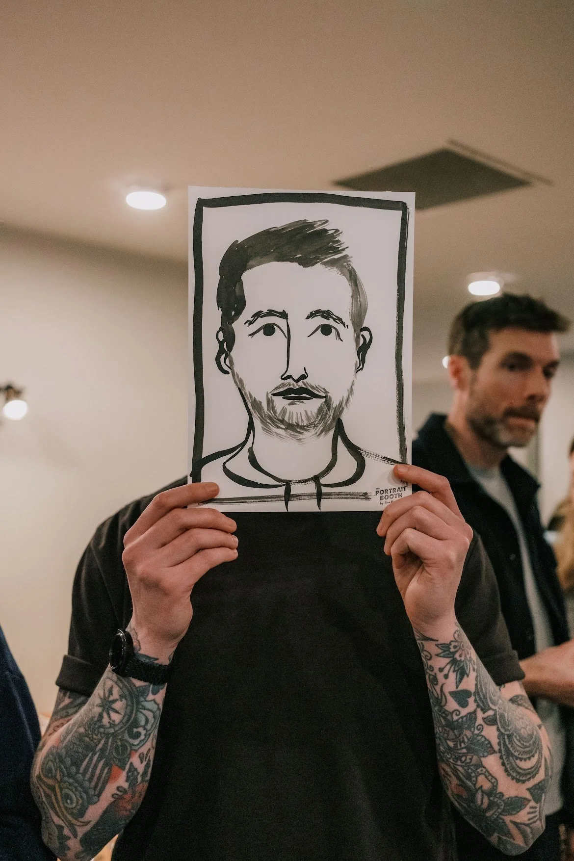 A guest shows off their finished Rubbish Portrait at an event, holding the playful black-ink artwork in front of their face. The bold brushstrokes capture their features with Lucie Sheridan’s signature charm and humour, creating a one-of-a-kind keeps
