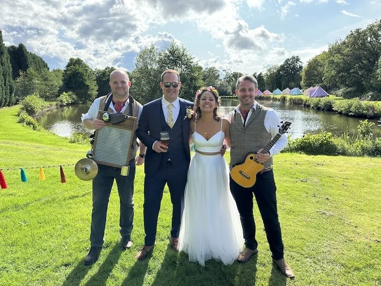The Ukes of Hazard smile with a newlywed couple at an outdoor summer wedding beside a scenic lake, complete with colourful tipi tents in the background. The band members wear tweed waistcoats and red neckerchiefs, holding their ukuleles and washboard