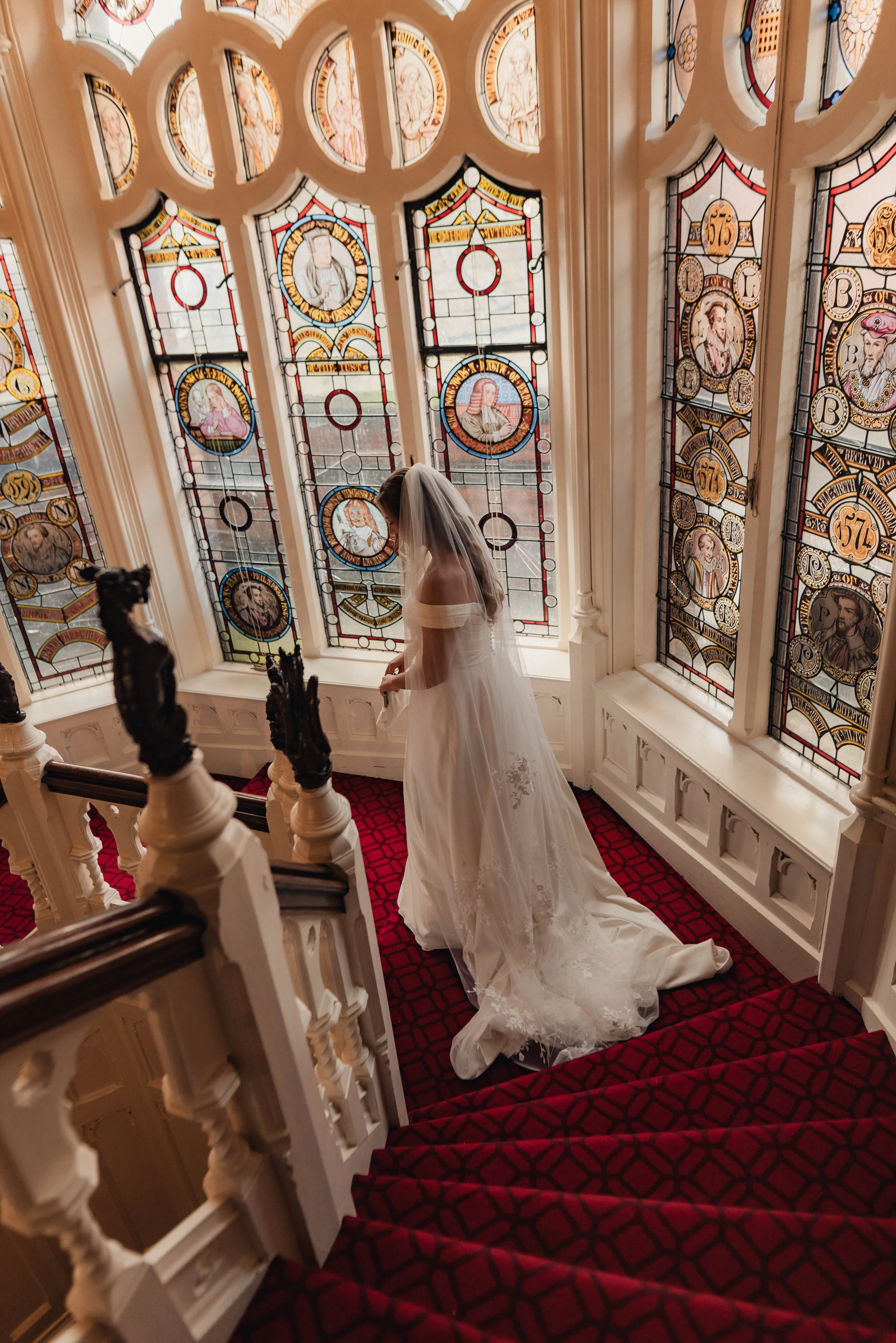 A bride in an elegant off-the-shoulder wedding gown descends a grand staircase lined with rich red carpet, framed by ornate stained-glass windows in a historic venue. Her long veil trails gracefully behind her as warm natural light highlights the int