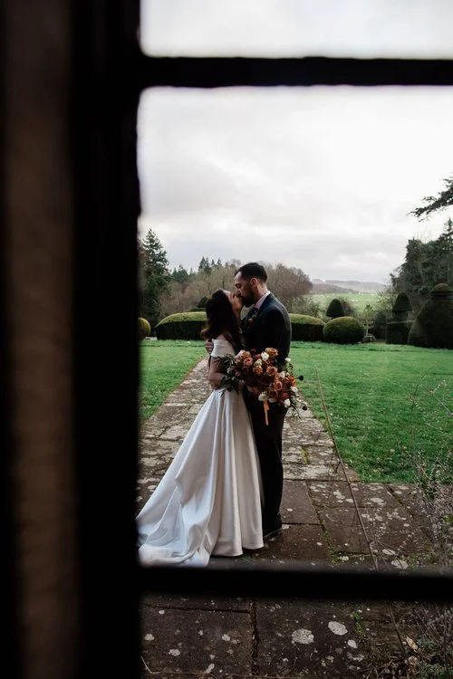 A beautifully intimate wedding portrait captured by Mirrorbox Photography, taken through a window frame to create a natural, documentary-style composition. The couple stand close together on a stone terrace overlooking manicured gardens and rolling c