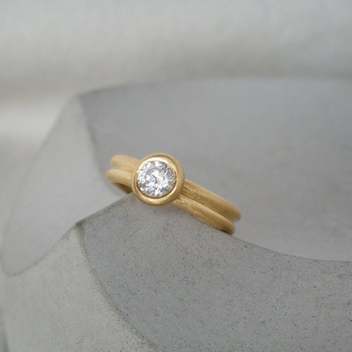 A minimalist Natalie Vernazza yellow gold engagement ring featuring a round brilliant cut diamond in a smooth bezel setting, paired with a delicate double band for a modern yet timeless look.