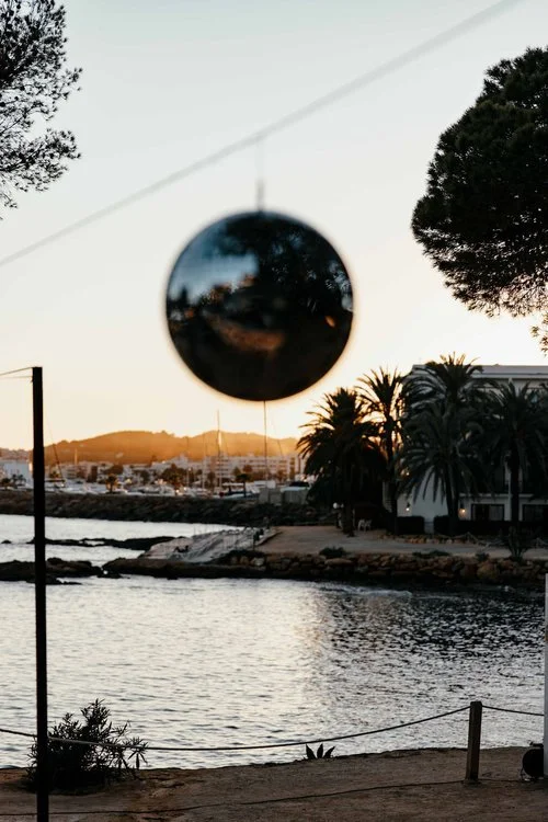 A disco ball hanging outdoors at sunset over a coastal wedding venue, with palm trees and yachts in the background. This romantic seaside setting creates the perfect backdrop for a wedding party, combining boho vibes with festival-style decor.