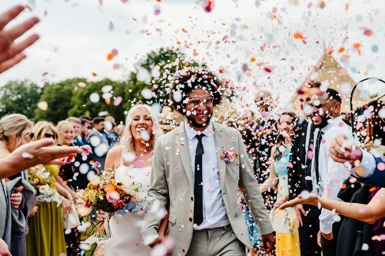 A burst of colourful confetti fills the air as the couple walk hand in hand through a cheering crowd with the groom smiling broadly beneath the fluttering pieces and the bride glowing beside him holding a bright bouquet while guests line the path tos