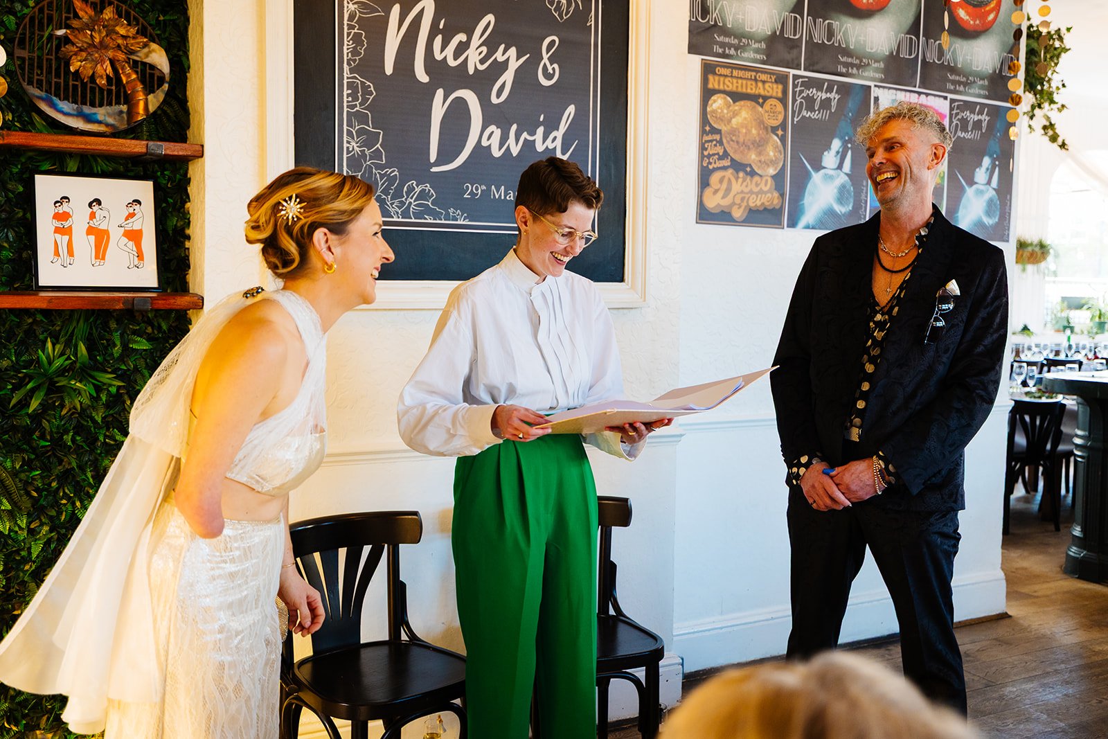 A joyful wedding ceremony captured indoors, with wedding celebrant Chloe Green leading a fun and heartfelt moment between a stylish bride in a shimmering gown and a groom in a chic black suit. The couple are laughing together as the celebrant reads f