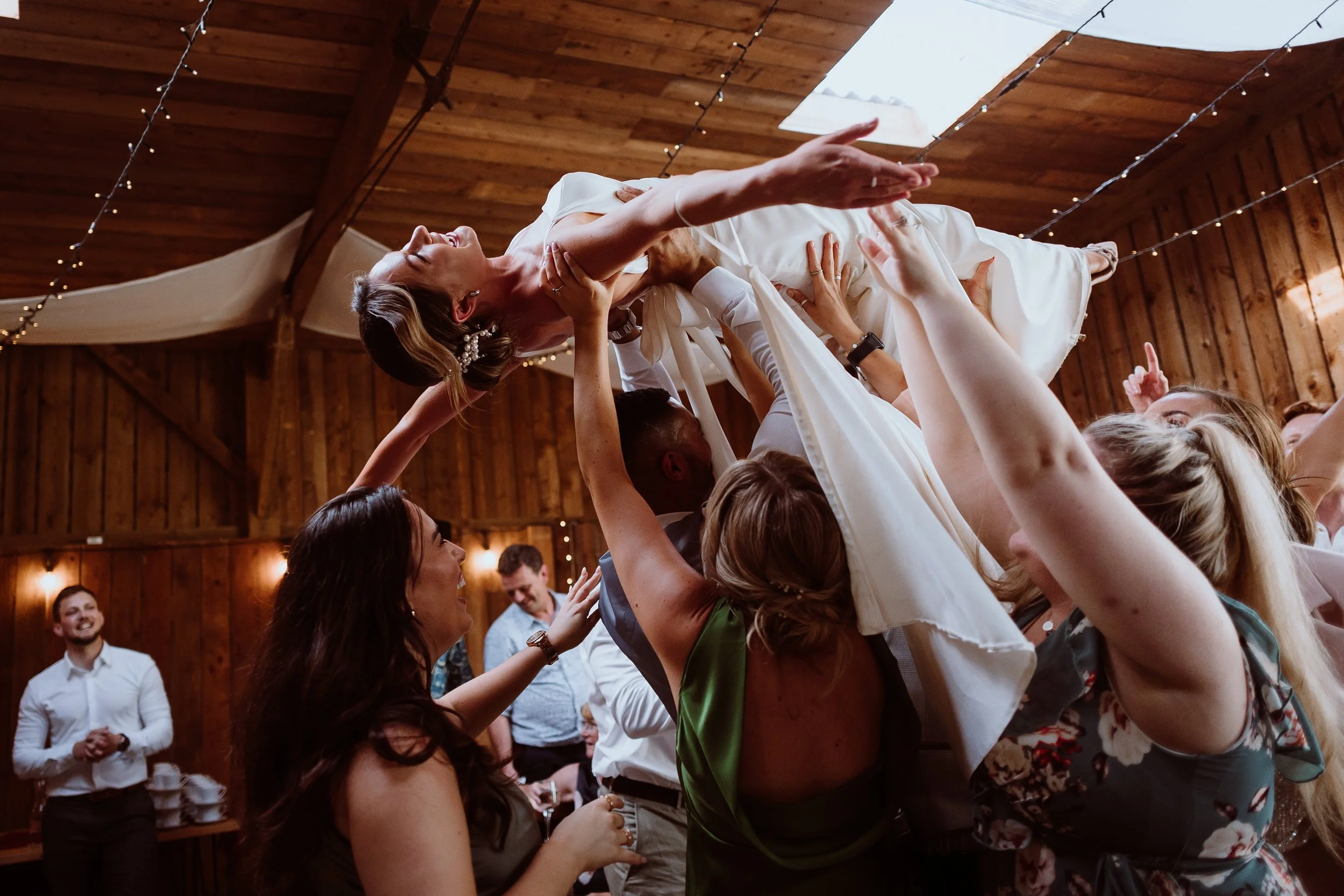 A wild and joy filled celebration moment captured by Joy Louise Photography, showing the bride being lifted high into the air by her guests on the dance floor. Her arms stretch wide as she laughs, surrounded by friends reaching up to support her bene