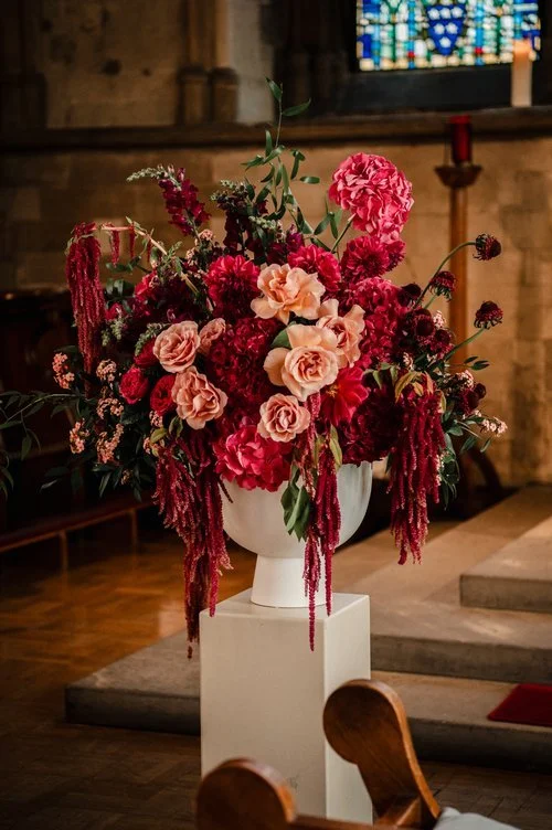 Dramatic church wedding floral arrangement by Botanique Workshop featuring rich red and deep pink blooms with soft peach roses, cascading amaranthus and natural greenery in a modern white urn. Styled within a historic ceremony setting, this bold and 