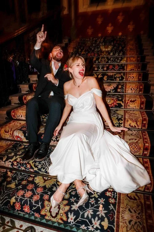 A fun and expressive wedding moment captured by Amy Faith Photography, showing the couple laughing together as they sit on the patterned staircase of their venue. The bride’s off-the-shoulder gown flows around her in soft folds while her bright lipst