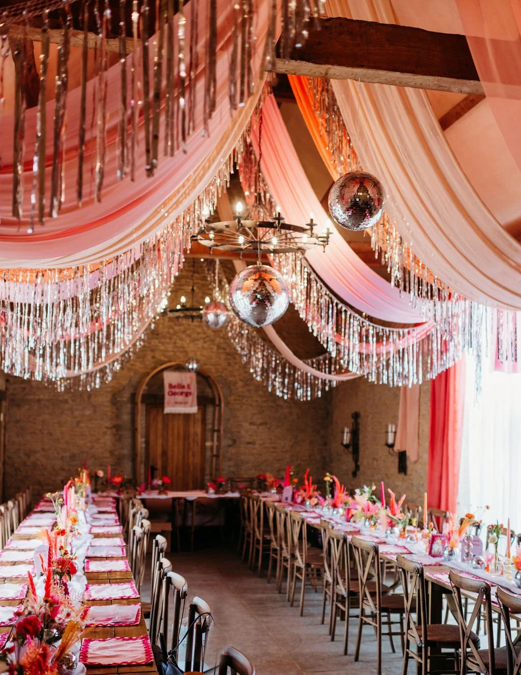 Vibrant wedding reception by AMW Events, featuring long banquet tables decorated with bright florals, overhead draped pink and orange fabrics, sparkling tinsel garlands, and disco balls hanging from a rustic chandelier.