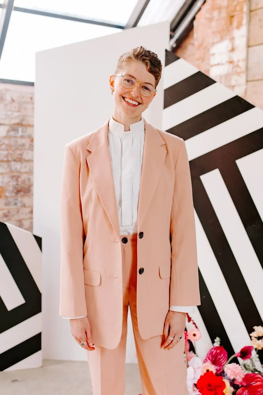 A modern UK wedding celebrant smiles warmly while standing in a stylish industrial venue decorated with bold monochrome patterns and bright floral accents. Dressed in a tailored peach suit and crisp white shirt, the celebrant exudes professionalism a