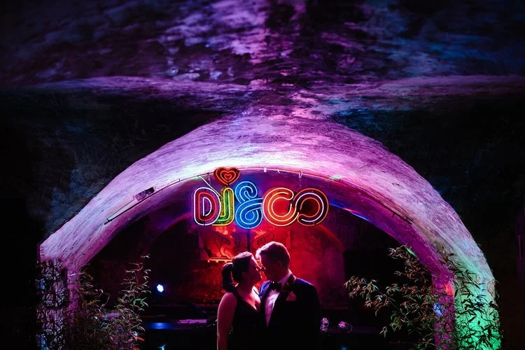 A couple stand close together beneath a glowing neon “DISCO” sign in an atmospheric underground space filled with vivid purples, pinks, and greens. Their silhouettes are softly lit by the colourful lights as they share an intimate moment before the d