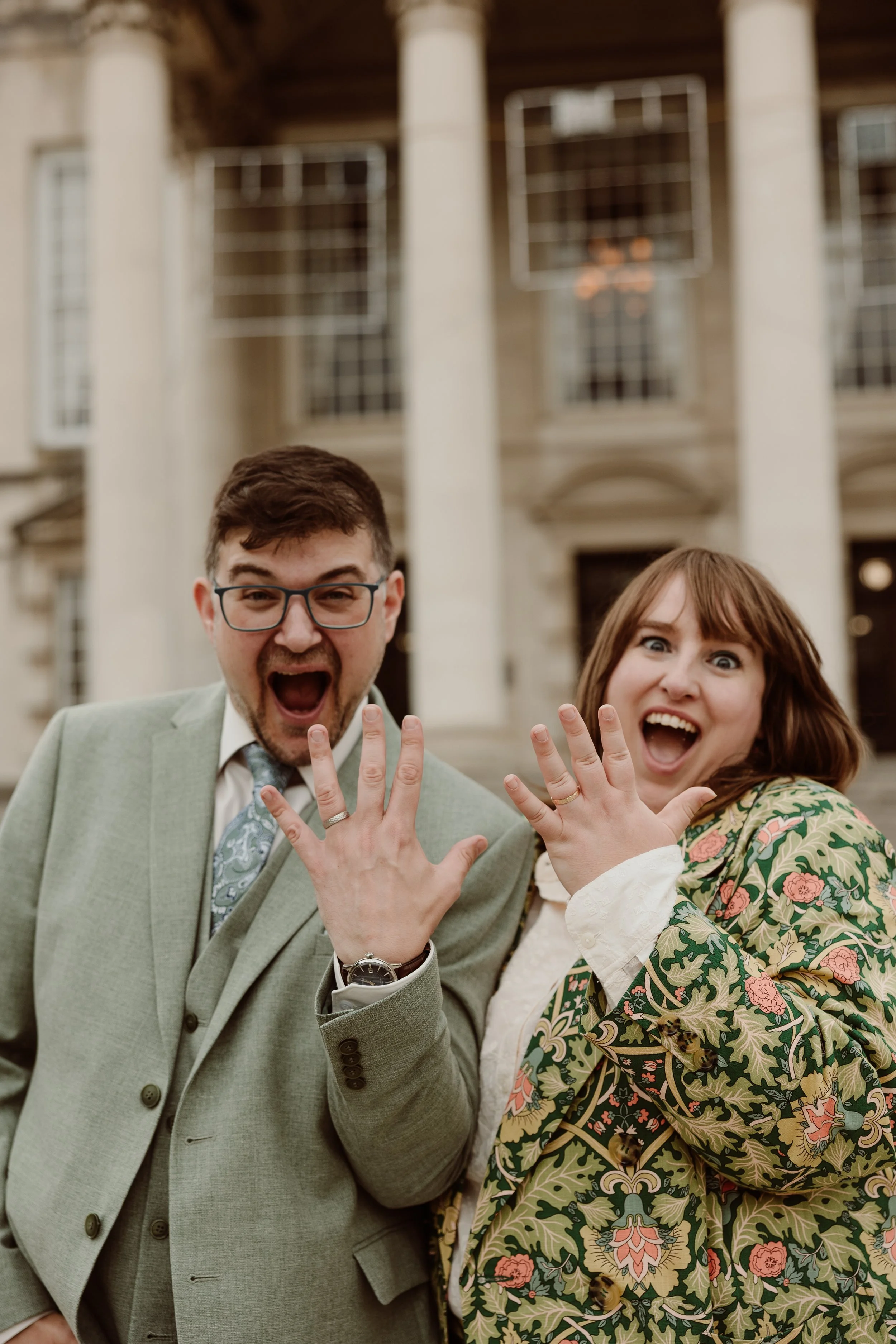 A fun and expressive newlywed portrait captured by Joy Louise Photography, showing the couple proudly displaying their wedding rings with huge, joyful surprise on their faces. Standing in front of grand architectural columns, they hold their hands ou