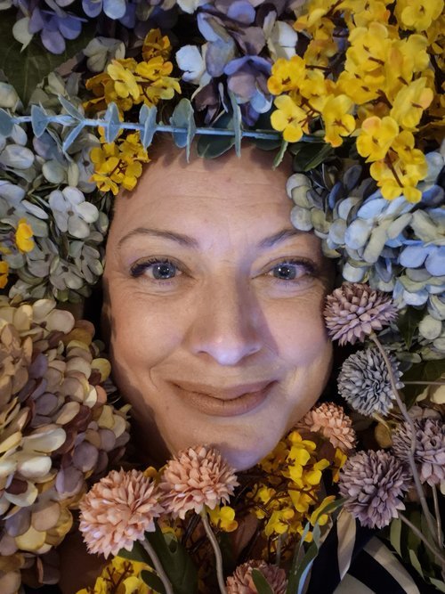 Close-up portrait of Melly the Celebrant surrounded by a vibrant arrangement of colourful flowers in soft pink, yellow, lavender, and blue tones. The floral frame creates a whimsical, artistic composition that highlights natural beauty and creativity