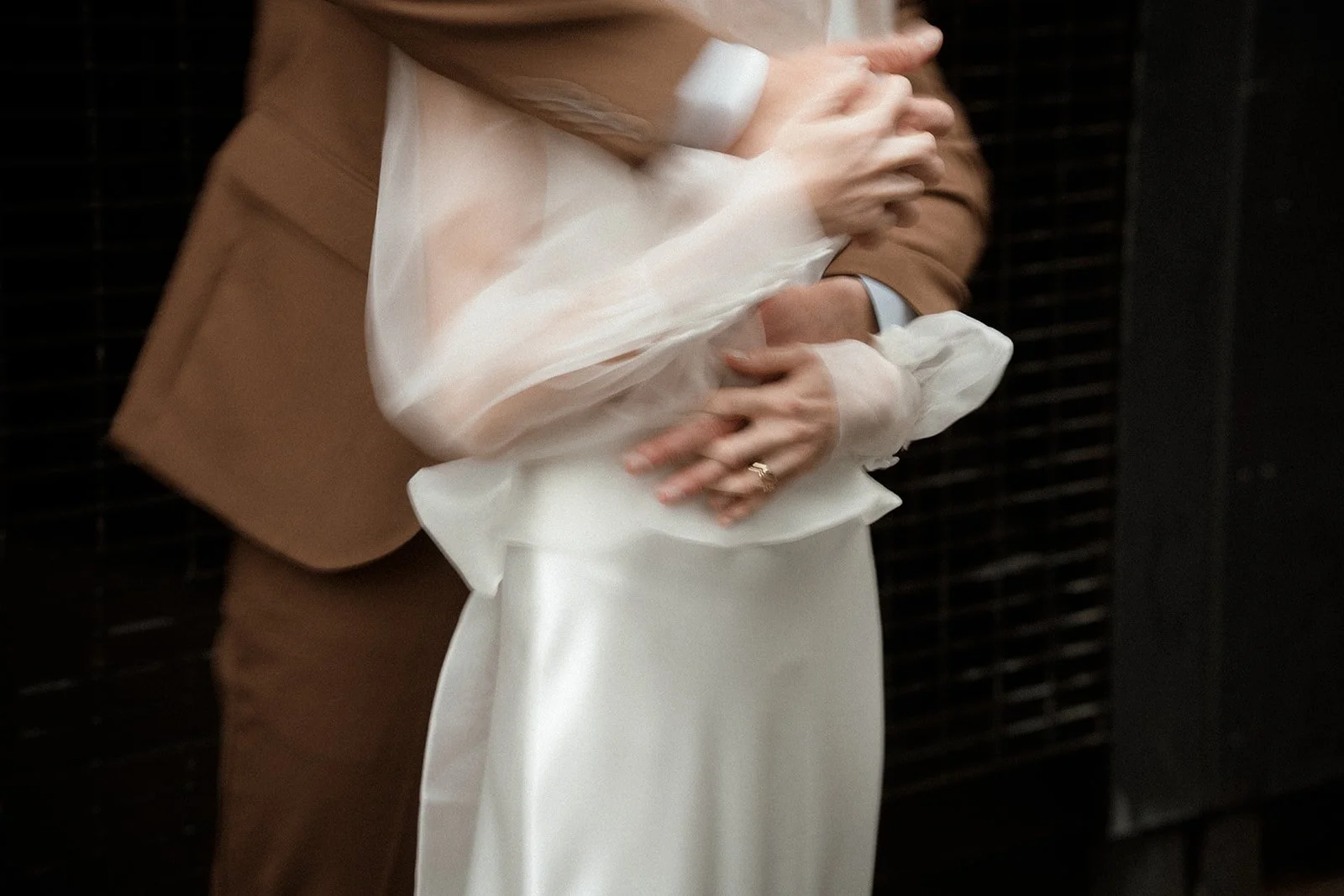A close up wedding portrait shows a groom in a brown suit embracing his bride who is wearing a white dress with sheer, billowing sleeves, as their hands overlap at her waist while standing against a dark textured wall, highlighting the movement of th