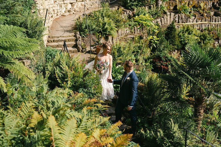 A newly married couple walk together through a sunlit garden filled with lush ferns and tropical greenery, beautifully captured by Christy Photography. The stone pathways, natural textures and bright foliage create a stunning backdrop that highlights
