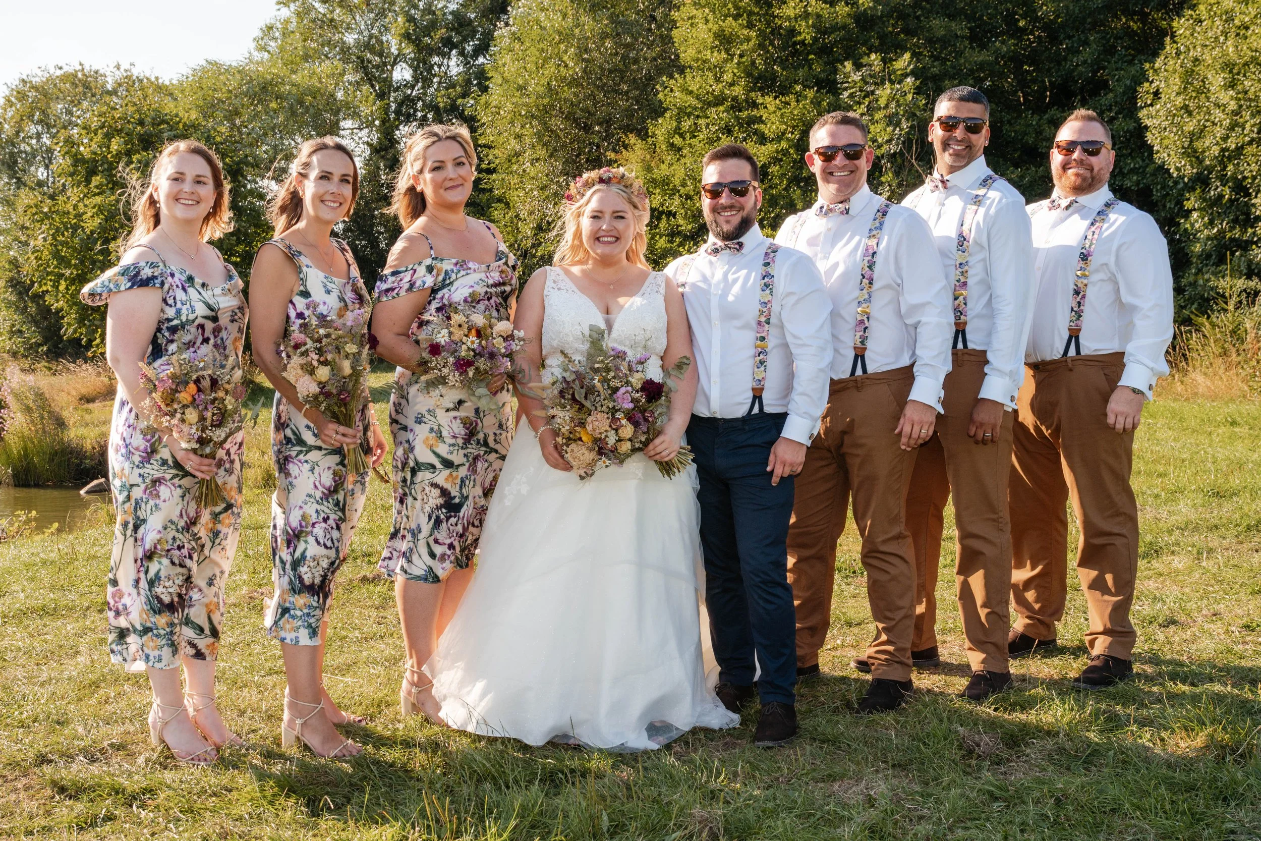 A wedding party stands on sunlit grass with trees behind them, the bride in a white gown and flower crown holding a mixed dried bouquet, flanked by bridesmaids in matching floral dresses carrying similar bouquets, while the groomsmen wear white shirt