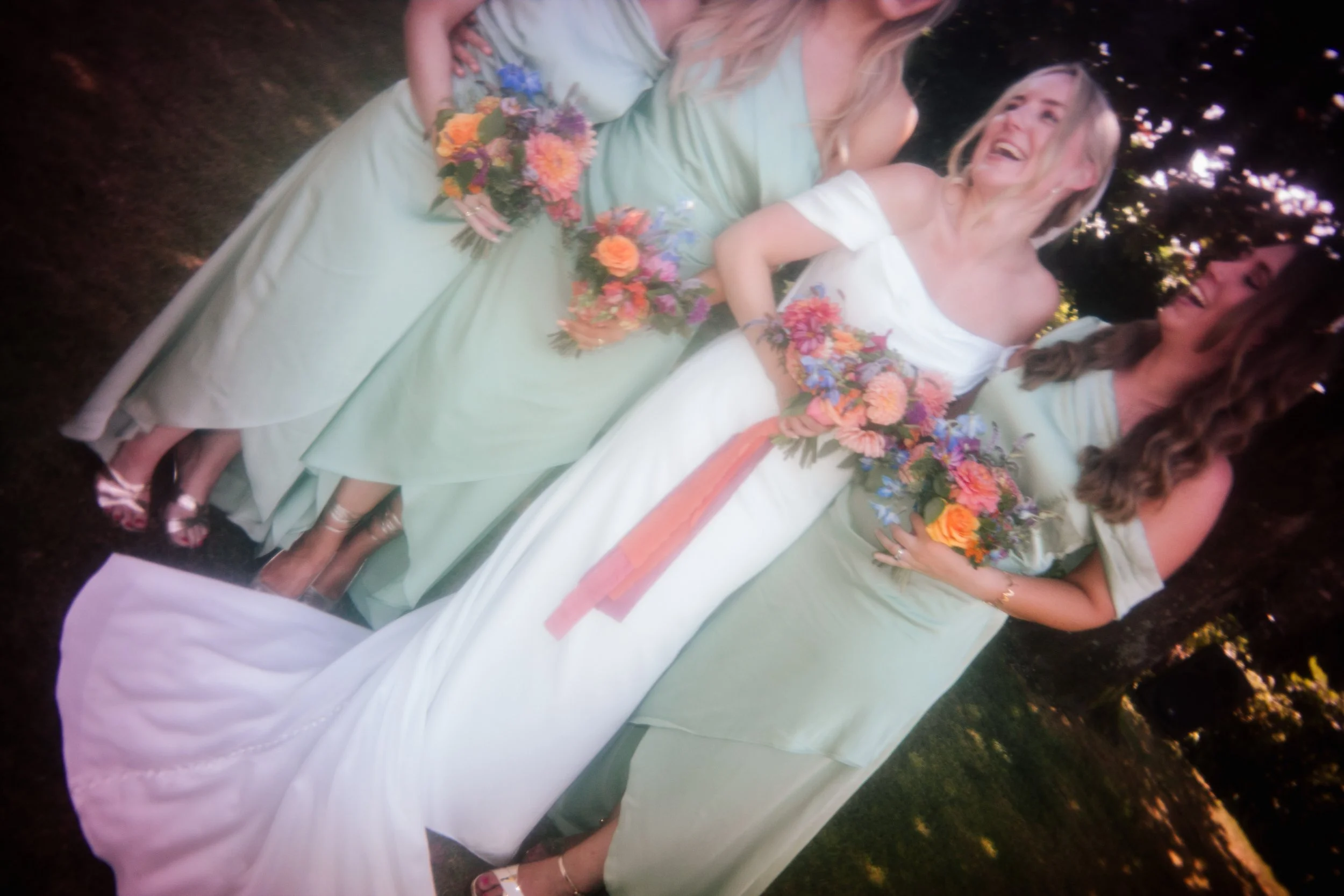 Dreamy, soft-focus wedding party portrait showing a bride in an off-the-shoulder white gown laughing with her bridesmaids in sage green dresses, each holding colourful summer bouquets. Captured outdoors under dappled shade, this candid film-style sho