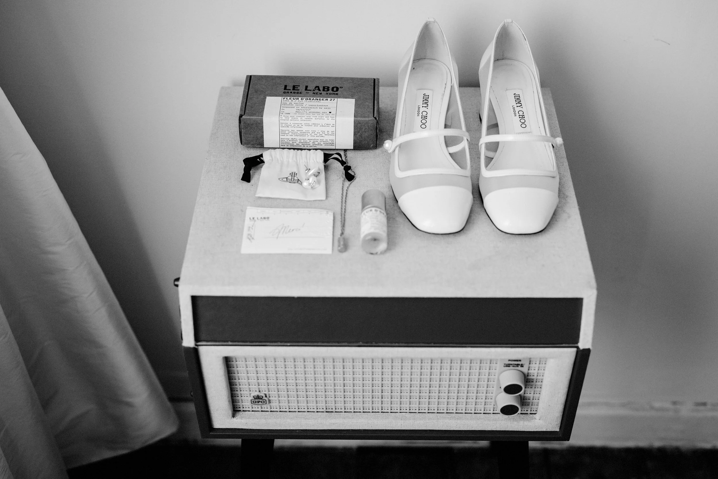 An elegant black and white wedding detail captured by Teddy Photo and Film showcases carefully arranged bridal accessories, including classic Jimmy Choo wedding shoes, Le Labo fragrance, jewellery and keepsakes displayed on a vintage bedside cabinet,