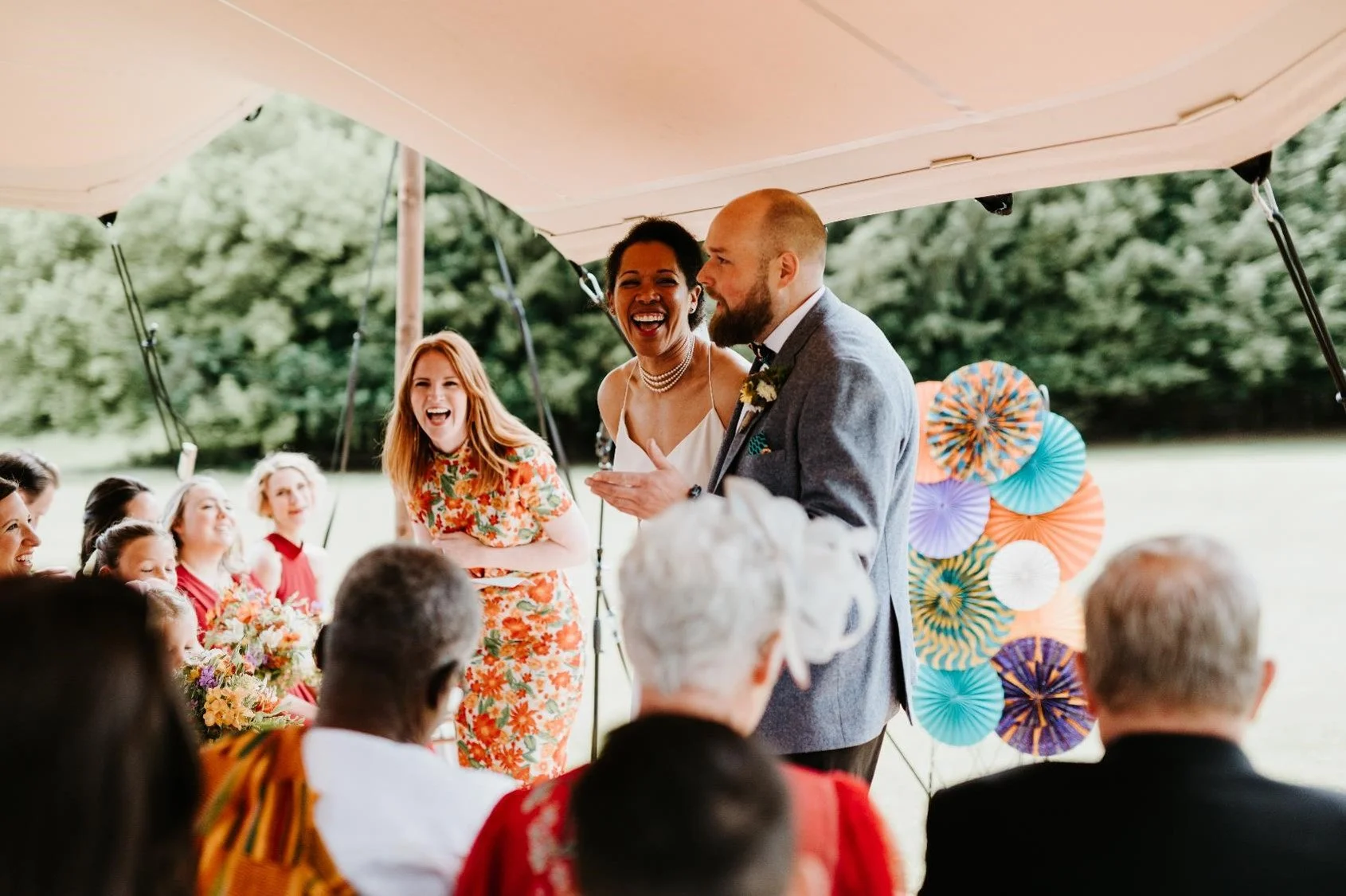 Joyful outdoor wedding ceremony led by celebrant Meg Senior, dressed in a bright floral outfit, laughing alongside the couple under a canopy. The bride and groom share a lighthearted moment surrounded by smiling guests and colourful paper decorations