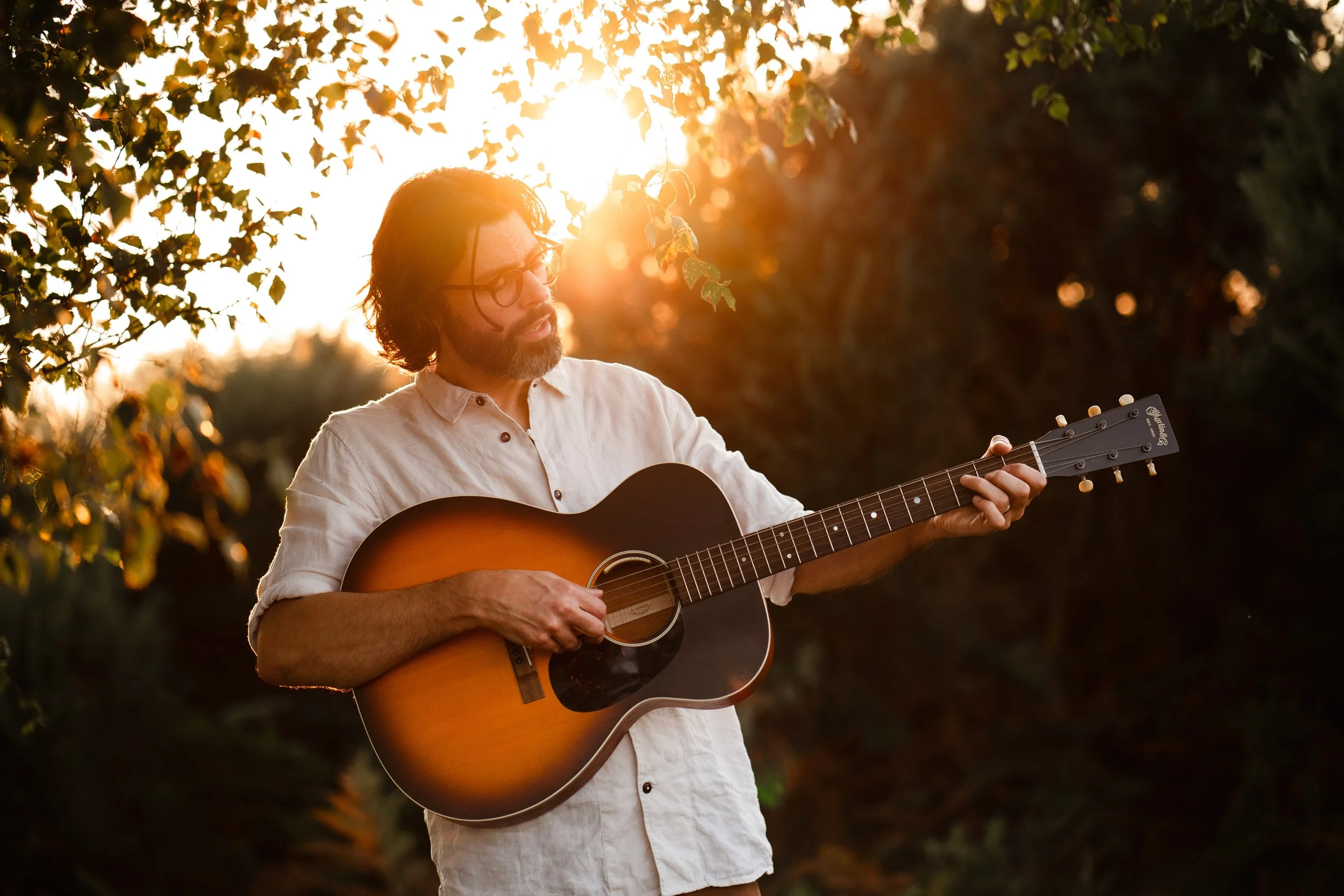 Thomas J Wilman performs outdoors at sunset, playing acoustic guitar beneath golden light. His relaxed and soulful style makes him a perfect choice for couples seeking romantic live music for their wedding ceremony or drinks reception. Clarkie Photog