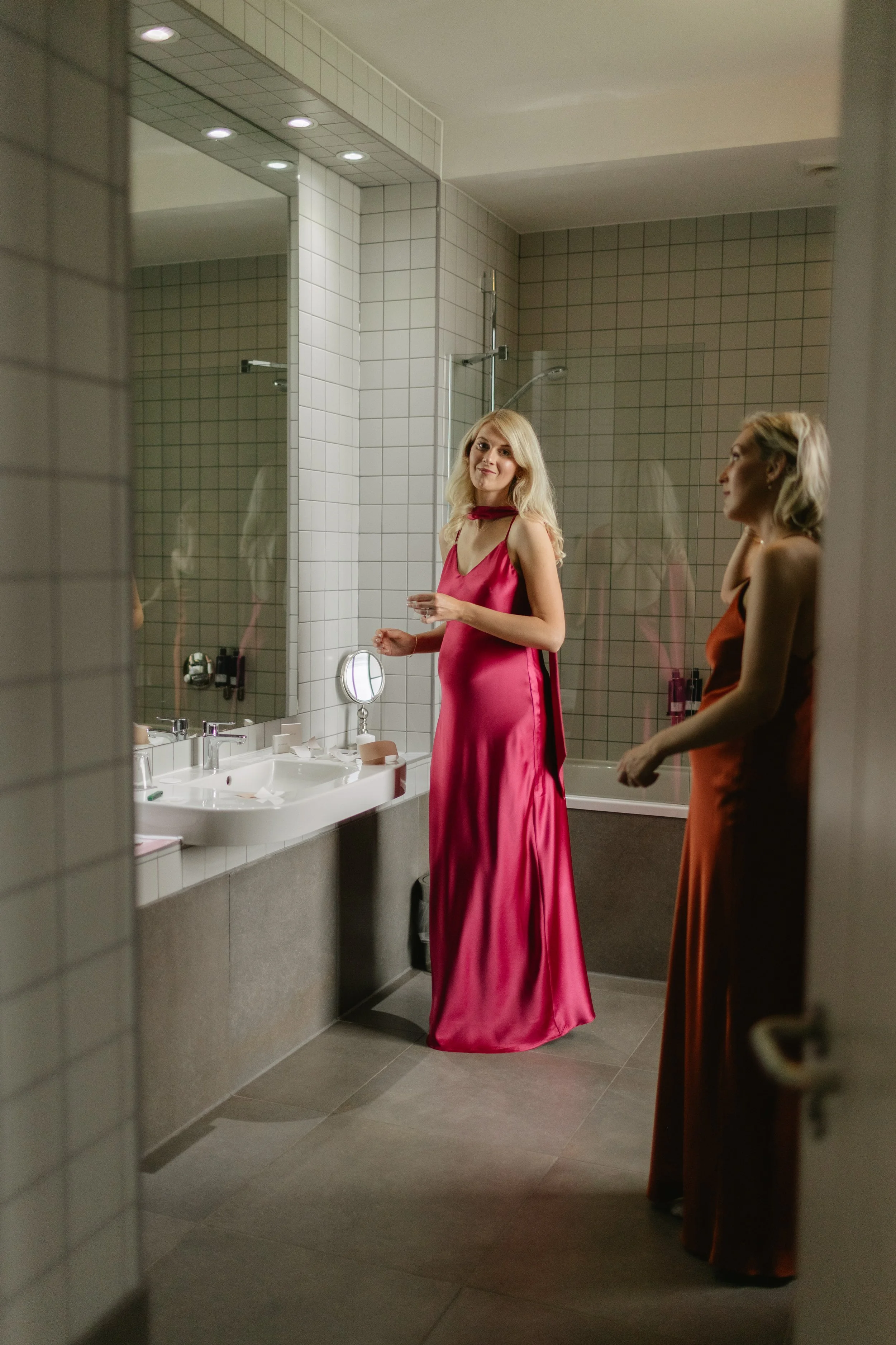 This image beautifully showcases Rewritten Bridal Wear’s modern satin bridesmaid dresses, known for their fluid drape and sophisticated silhouettes. The woman in the foreground wears a vibrant fuchsia gown with a bias-cut fit, cowl neckline, and long