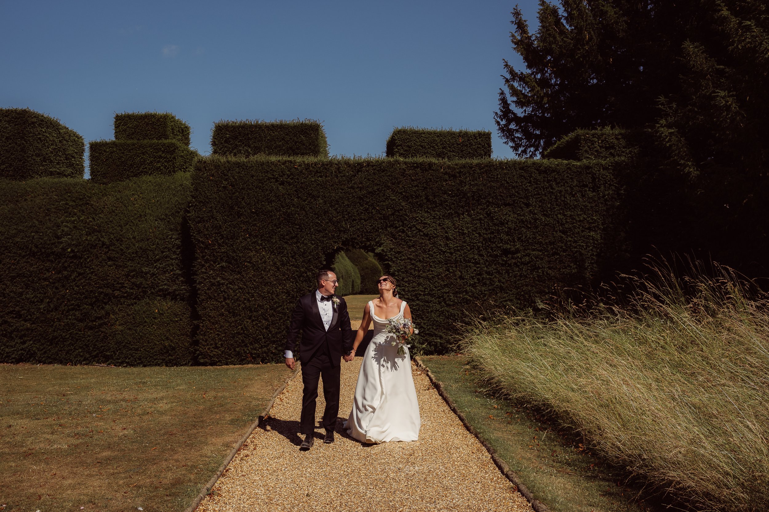 A bride and groom walk hand in hand along a sunlit gravel path in a beautifully manicured garden. Behind them stands an impressive wall of sculpted hedges with a circular archway, framed by clear blue sky and tall trees. The bride wears a classic whi
