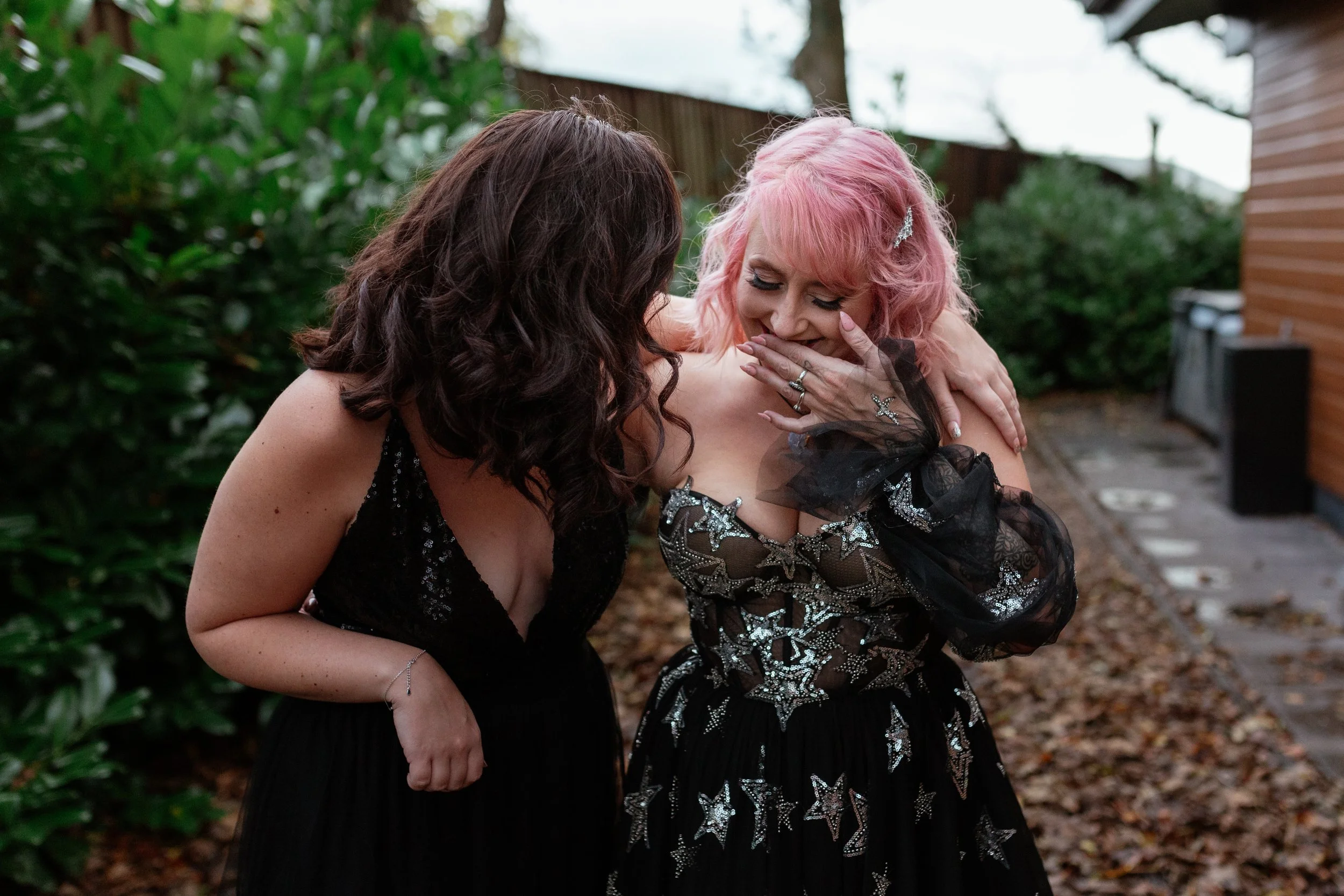 A heartfelt candid moment captured by Laura Wilson Photography, showing two women in elegant black dresses sharing a quiet laugh together. One woman with soft curls leans in closely while the other, with pastel pink hair and a star embellished gown, 