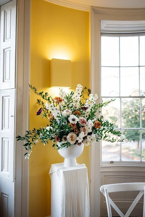 Elegant wedding floral arrangement by Botanique Workshop featuring white anemones, garden roses, delphiniums and abundant seasonal foliage displayed in a classic white urn. Positioned in a bright Georgian interior with yellow walls and large sash win