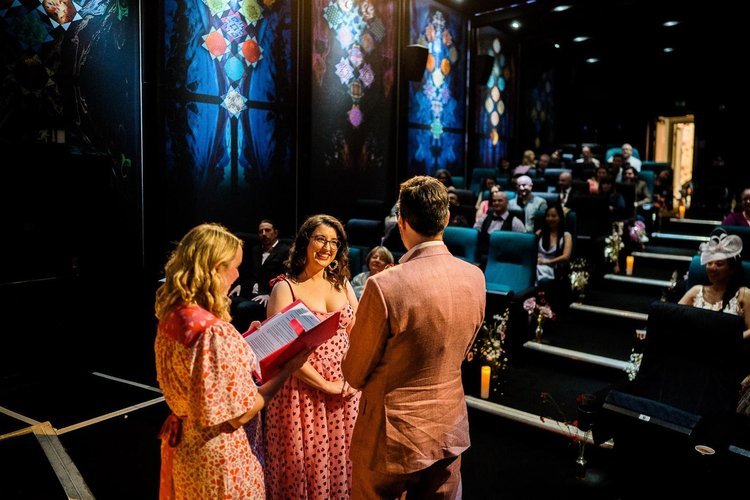 A fun and heartfelt wedding ceremony taking place in a colourful cinema venue, led by celebrant Frances Celebrates You in a bright floral dress. The couple, dressed in pink tones, share a joyful moment surrounded by smiling guests seated in theatre-s
