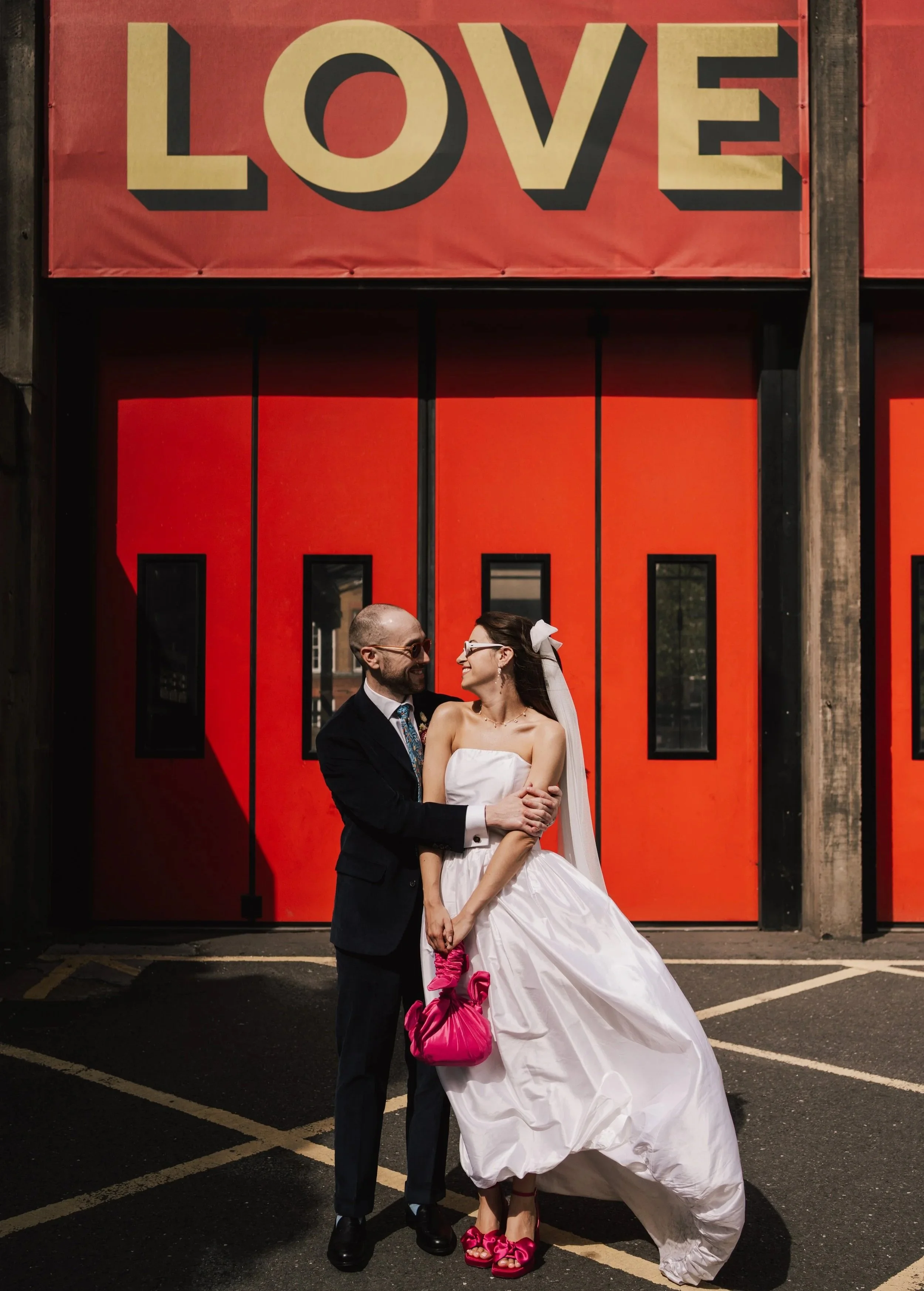 A modern couple standing in front of a red fire station with the word LOVE in bright yellow above them.