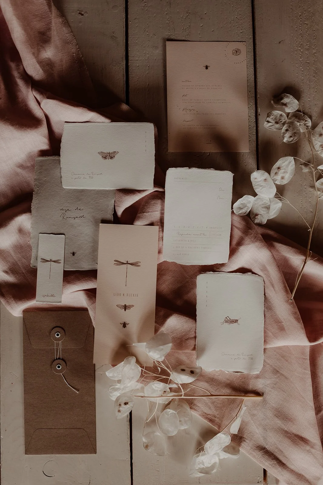 A romantic fine art wedding stationery flatlay featuring textured deckle edged cards in soft neutral tones adorned with delicate botanical and insect illustrations arranged on a muted pink linen cloth with dried honesty stems and a kraft string-tie e