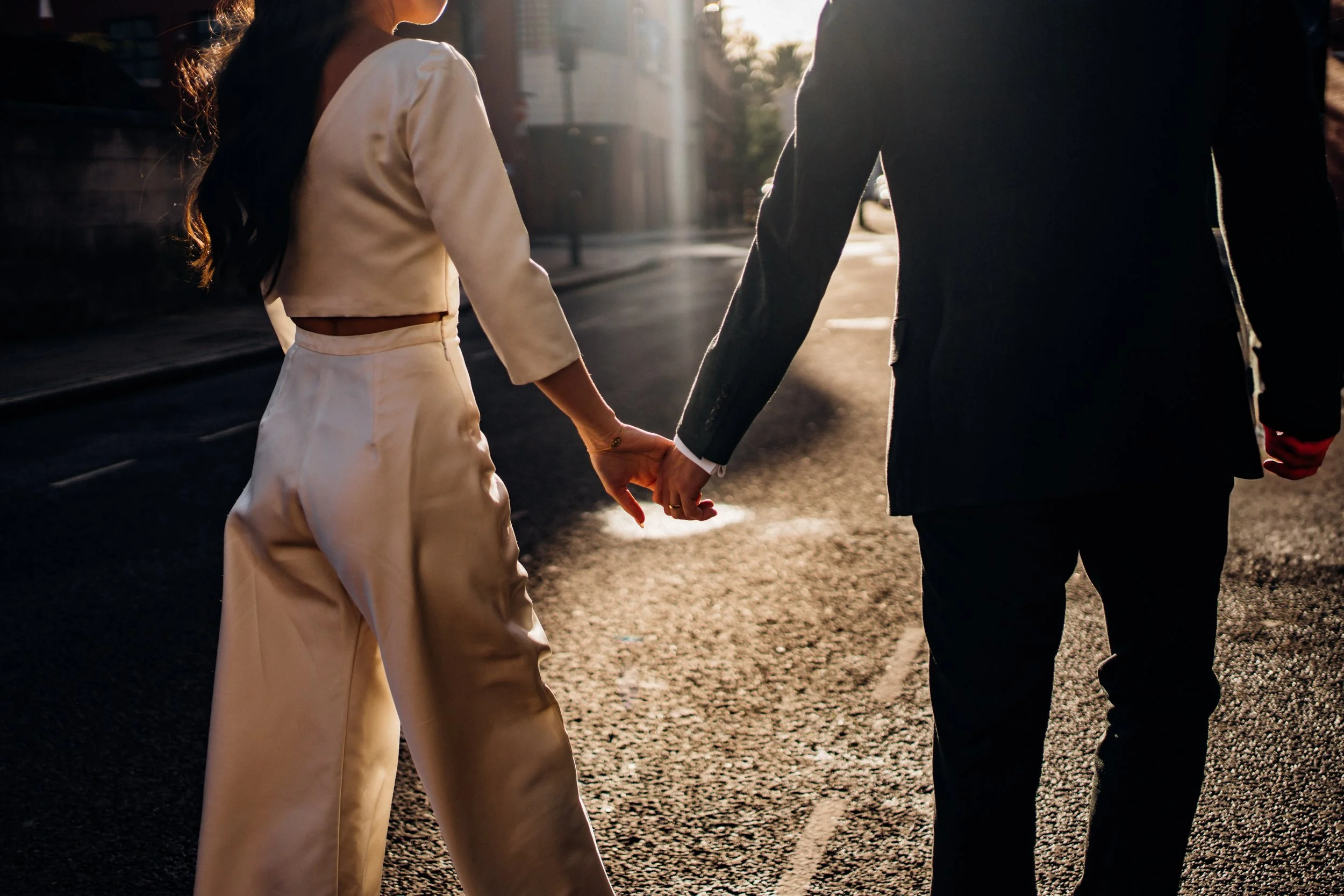 The image shows a couple walking hand in hand along a sunlit street with their backs to the camera, the low golden light creating a warm glow around them. The woman is wearing an ivory bridal two piece outfit with a cropped long sleeve top featuring 