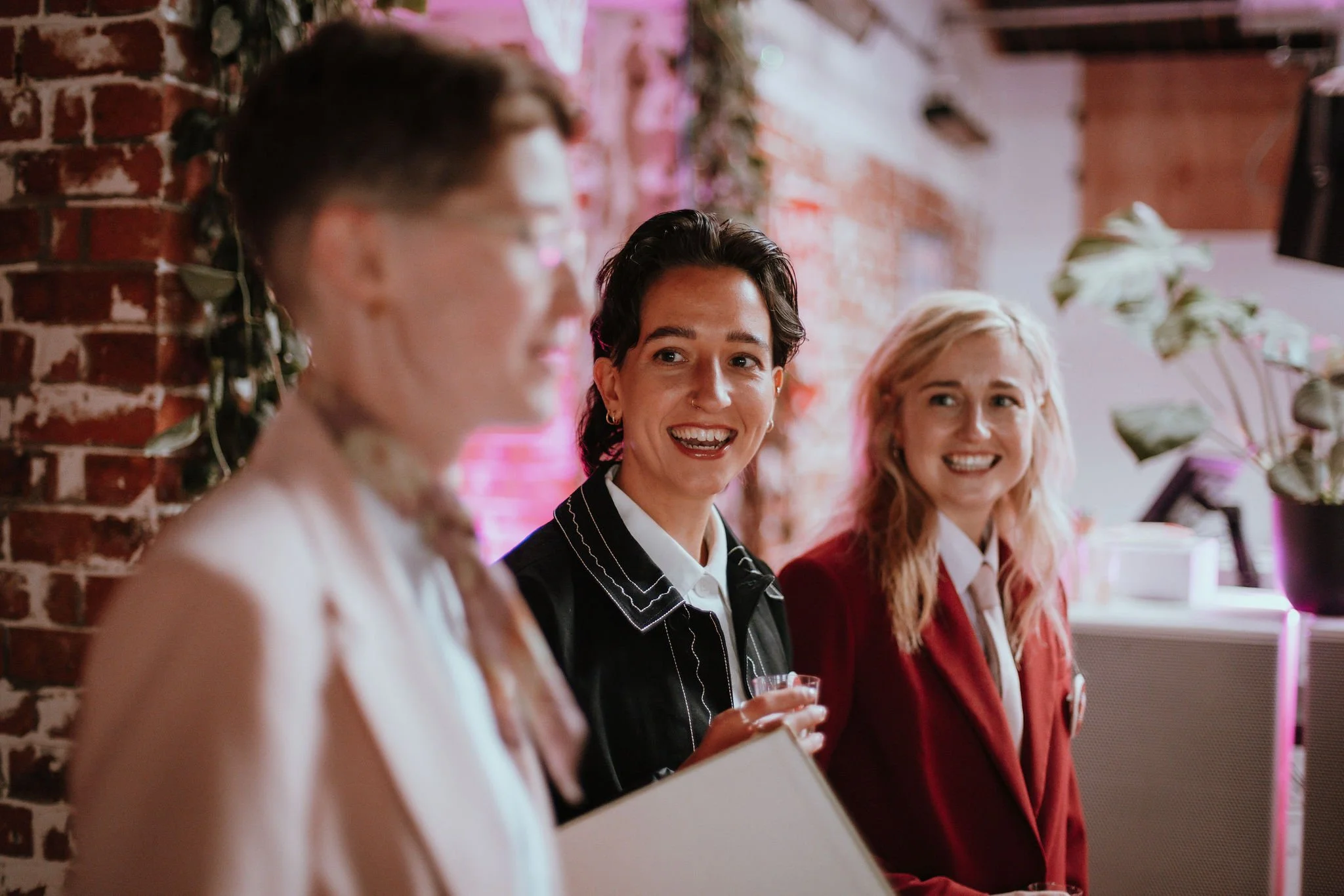 A nice moment at a UK wedding celebration featuring modern wedding celebrant Chloe Green with two happy newlyweds in a stylish, urban venue. The couple, dressed in chic suits, smile warmly as they enjoy a relaxed and inclusive ceremony atmosphere. Pe