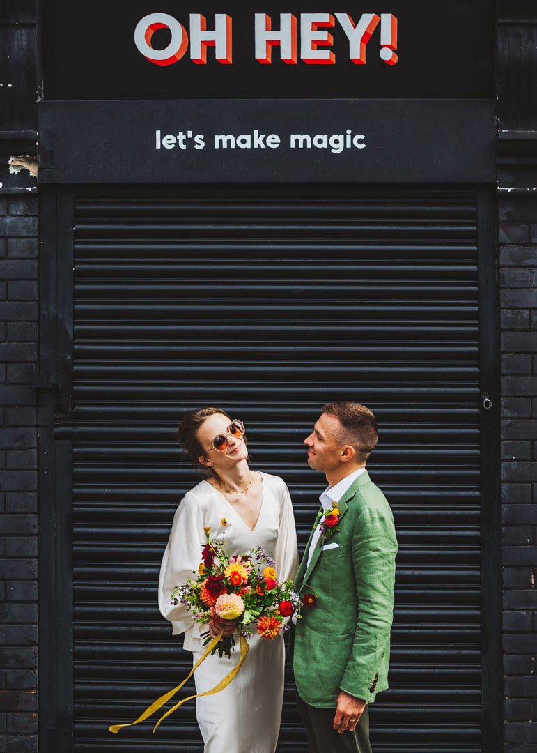 A cool and contemporary wedding portrait captured by Barbara K Photography, featuring a couple standing beneath a bold shopfront sign that reads “OH HEY! let’s make magic.” The bride wears stylish sunglasses and a minimalist white dress while holding