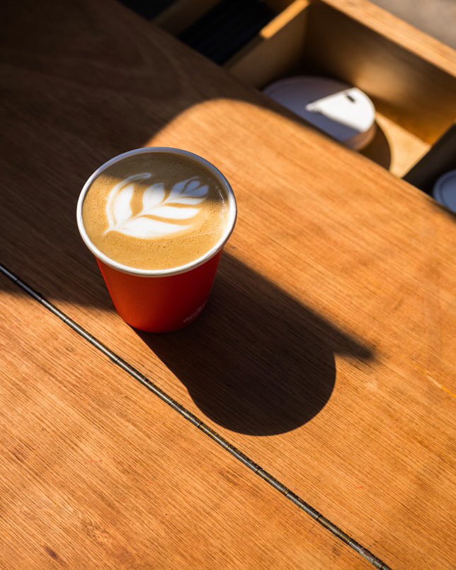 A warm close up capture from Red Panda Coffee showing a beautifully poured latte with delicate leaf latte art served in their signature red cup, resting on a handcrafted wooden counter in natural sunlight a cosy inviting detail perfect for showcasing