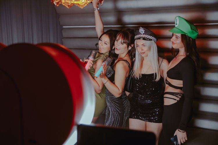 A group of friends poses at the Mark the Event photobooth, dressed for a night of celebration in stylish party outfits and fun props, including hats and microphones. The warm lighting and modern ring light setup create a glamorous backdrop for unforg