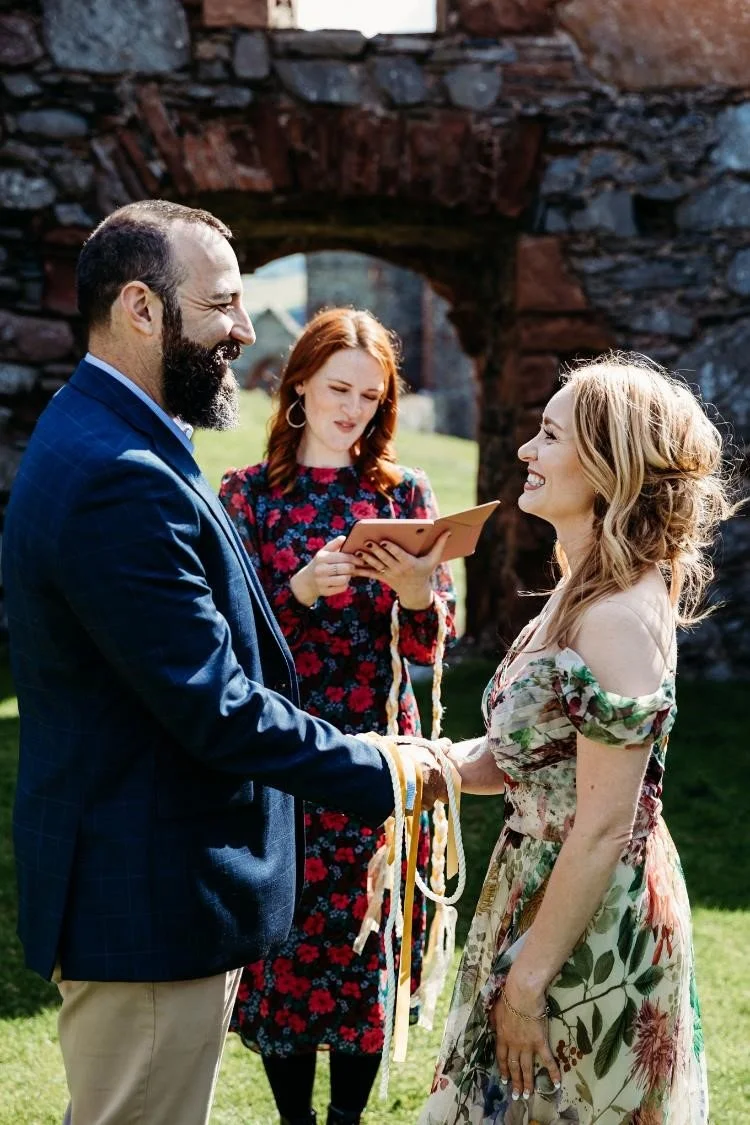 Wedding celebrant Meg Senior leading a hand-fasting ceremony for a smiling couple in a rustic outdoor setting. The bride wears a floral off-the-shoulder dress and the groom a blue checked jacket, as they exchange vows beneath the stone arch of ancien
