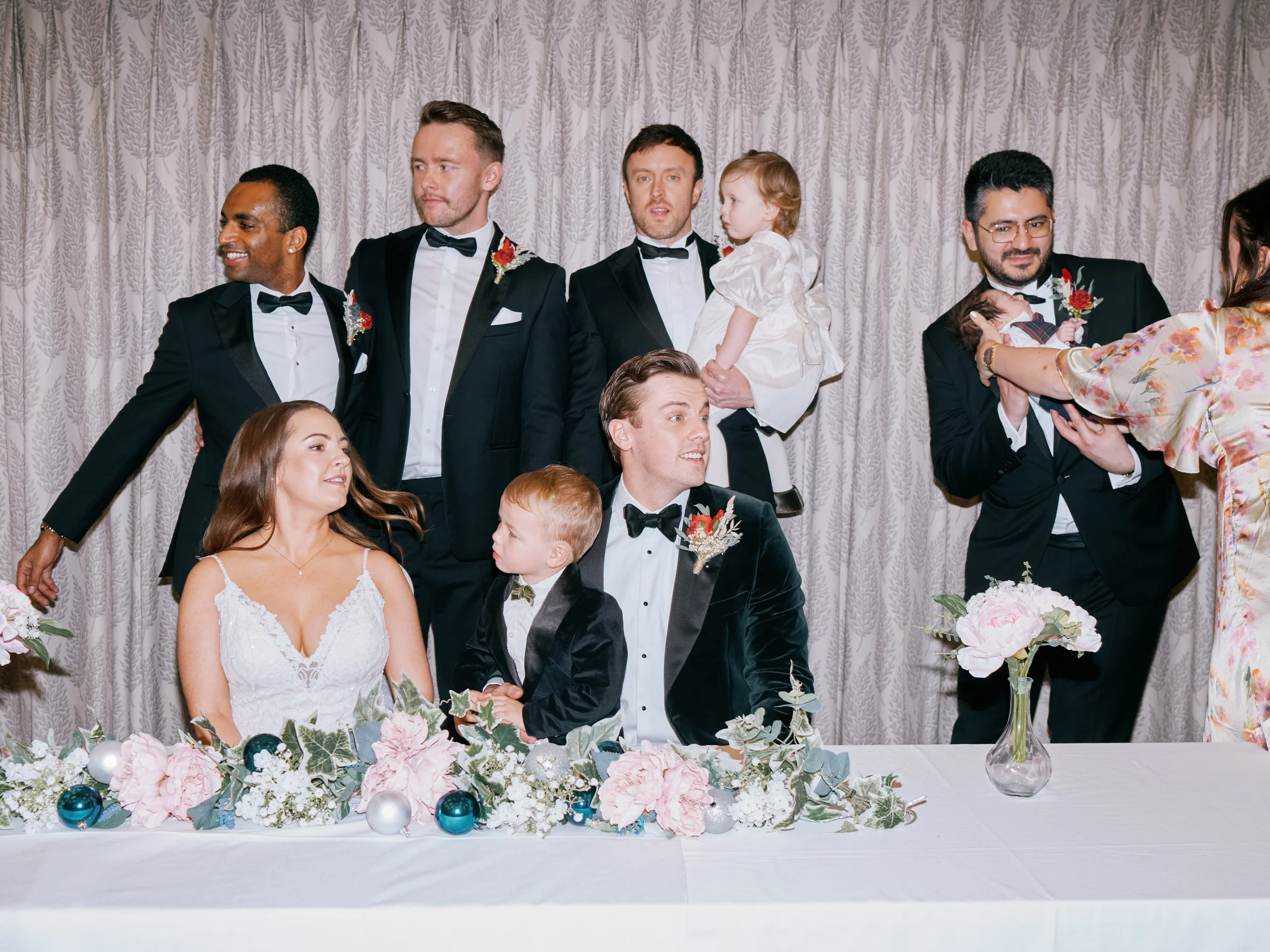 A lively and contemporary wedding portrait captured by Wayne La Photography, showing the couple seated at their signing table while surrounded by their bridal party and family in a beautifully candid moment. The bride’s delicate lace gown and the gro
