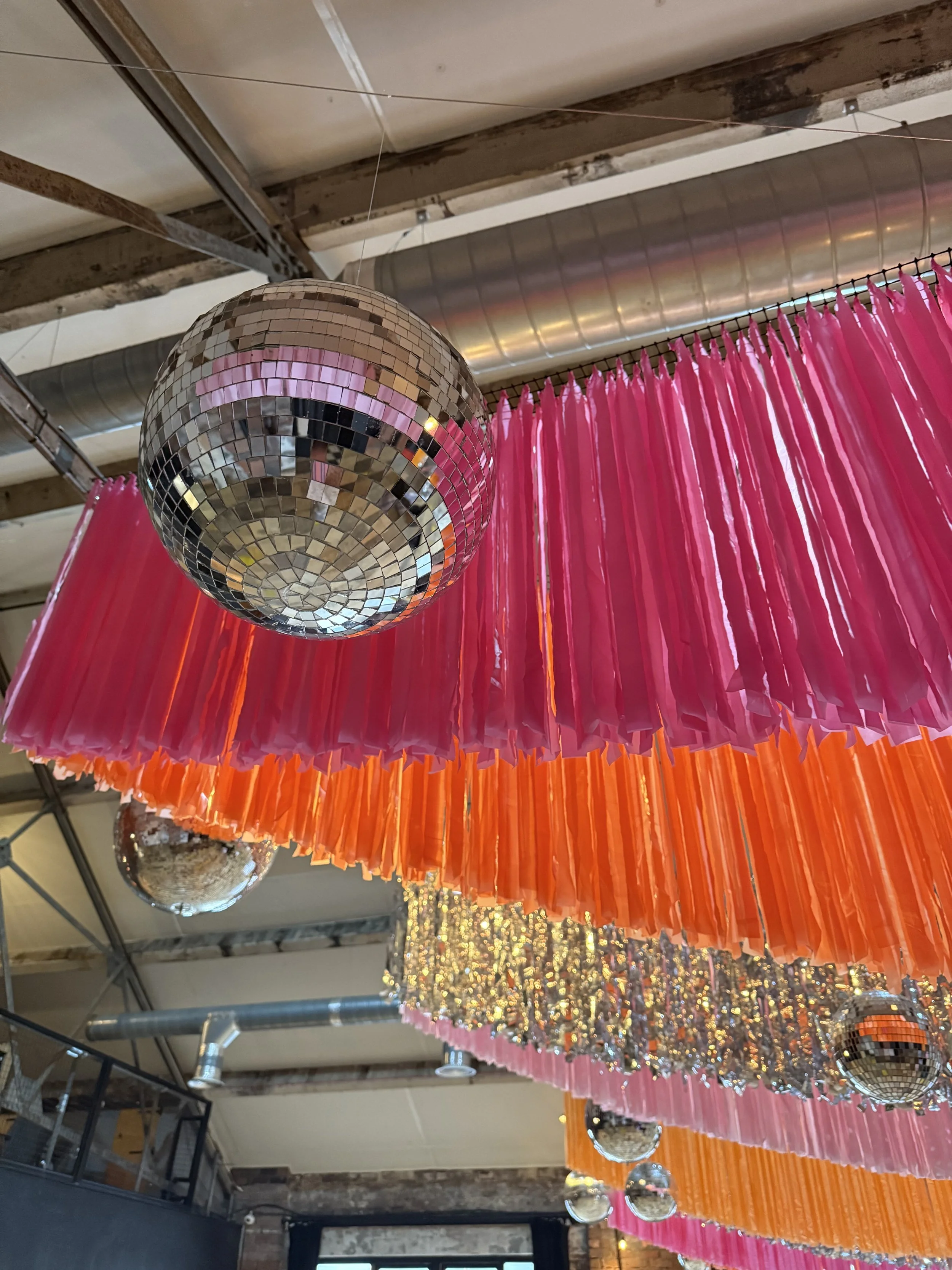 Hanging disco balls styled with pink and orange crepe streamers, creating a vibrant, retro-inspired ceiling installation. Colourful wedding decor hire ideal for modern, festival-style and alternative wedding venues.