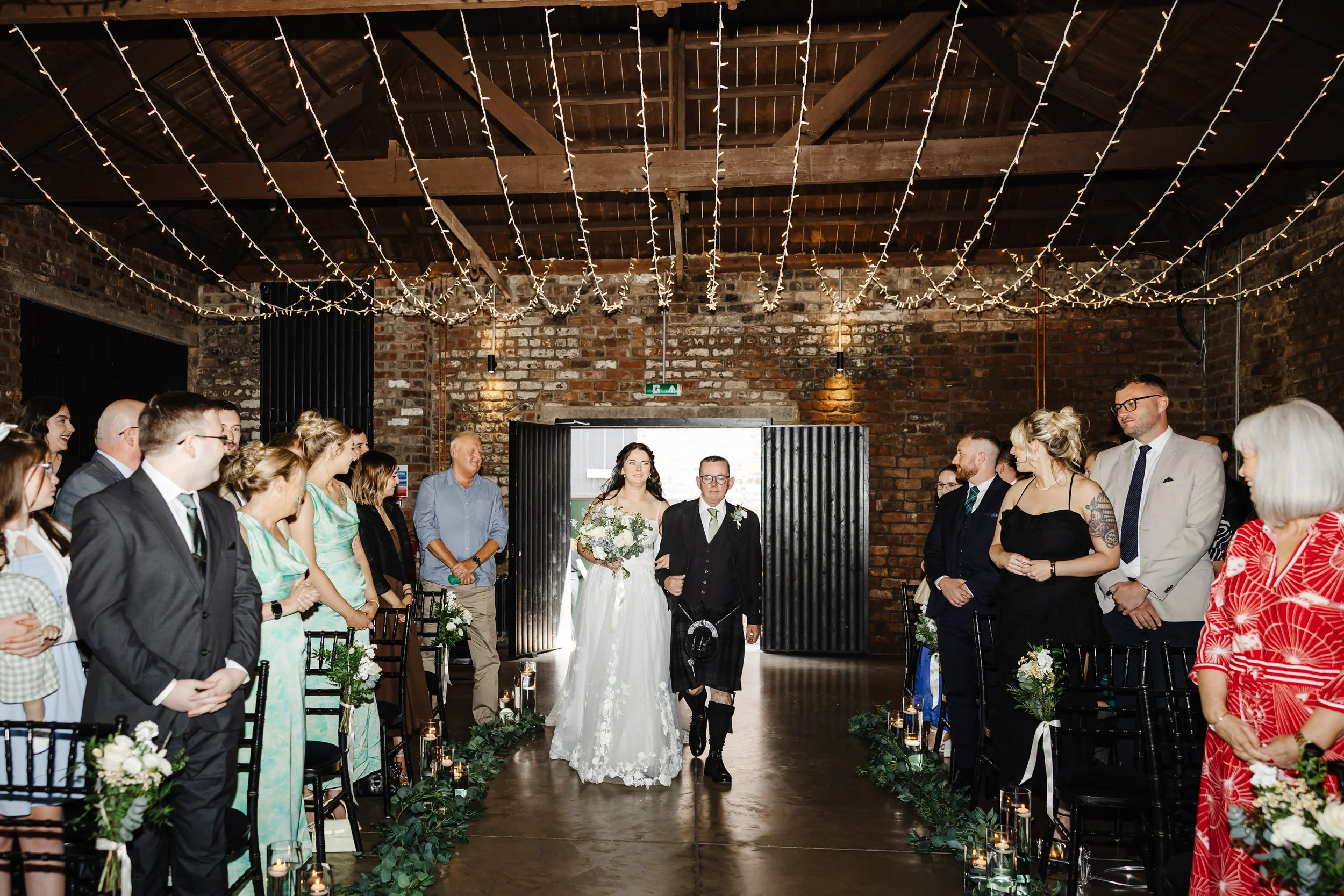Friends and family smile as Marie's father walks her down the aisle beneath string lights at The Engine Works.