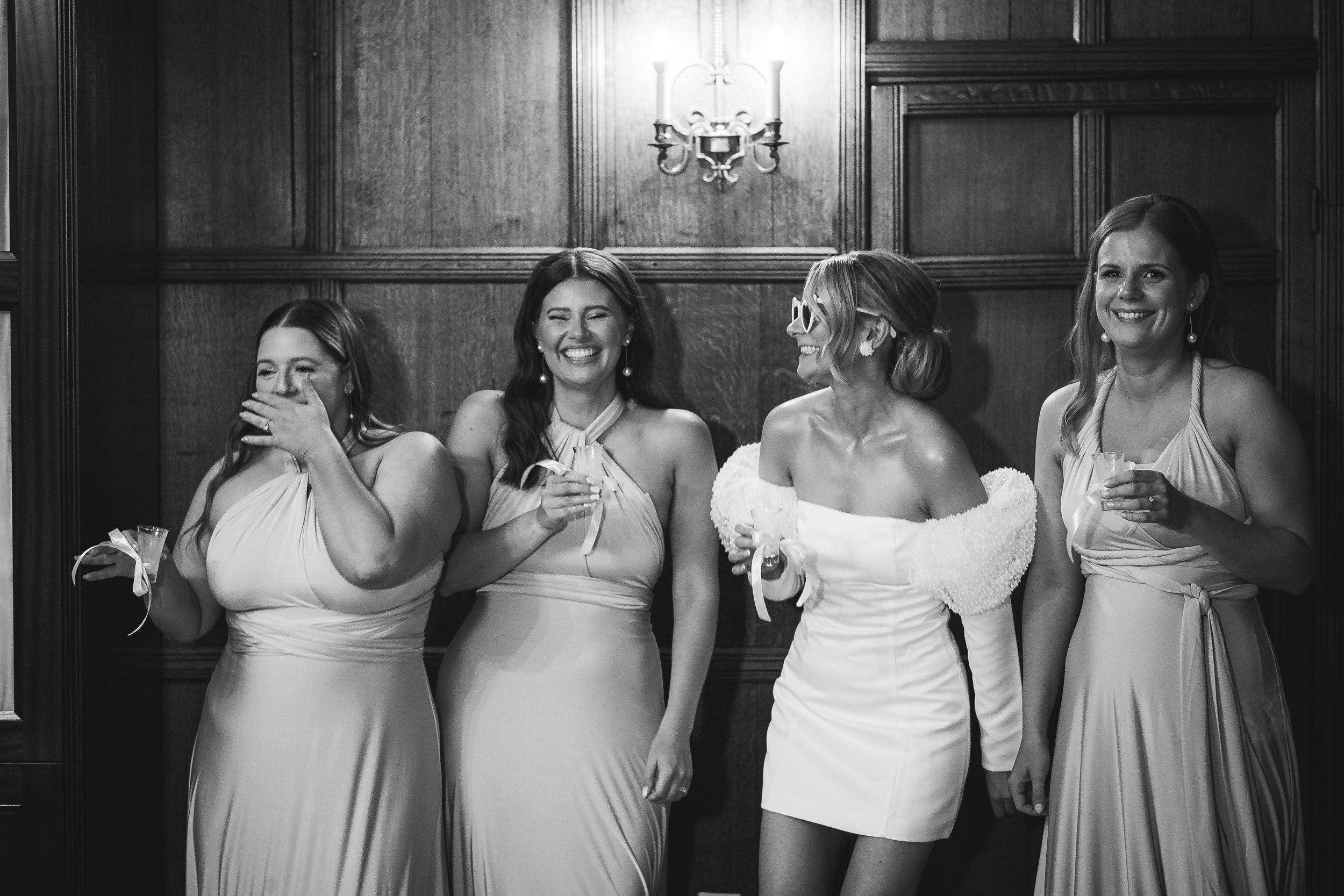 Four women stand together in front of dark wood paneling, all laughing and reacting with big, unfiltered emotion. Three of them wear matching light-colored dresses while the fourth, likely the bride, is dressed in a short, off-the-shoulder outfit wit