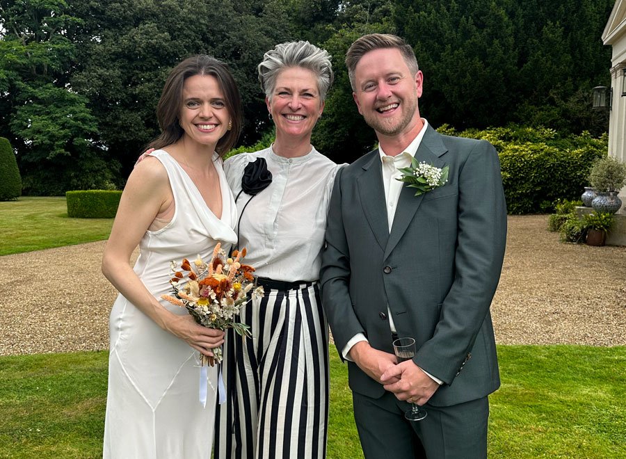 Three people stand close together on a lawn, smiling at the camera. The person on the left wears a sleeveless white dress and holds a small bouquet in warm, earthy tones, the person in the centre wears a light blouse with a dark flower detail and a b